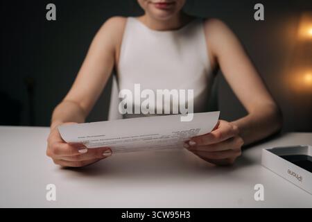 Professionelle Frau, die an einem minimalistischen Arbeitsplatz arbeitet und Schreibarbeiten am weißen Schreibtisch mit neuer ungepackter Smartphone-Kartonbox überprüft Stockfoto