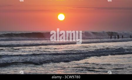 Lokale Fischer fischen im stürmischen Ozean während des farbenfrohen Sonnenuntergangs auf Bali und kreieren Silhouetten vor dem pulsierenden rosa Himmel mit riesigen Wellen im Vordergrund. Wunderschöner Natur Hintergrund Stockfoto
