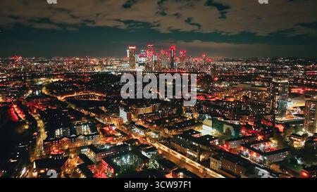 England, London: Die Wolkenkratzer der Canary Wharf und der umliegenden Wohngebiete leuchten nachts mit bunten Lichtern und bieten einen atemberaubenden Blick auf die Stadt von oben, von einer Drohne erfasst Stockfoto