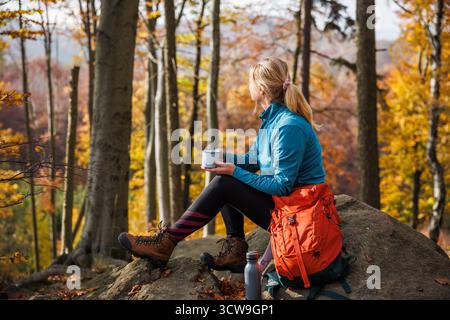 Frau, die sich nach erfolgreicher Wanderung ausruht. Weibliche Wanderer, die sich auf dem Felsen entspannen und im Herbstwald heißes Getränk trinken Stockfoto