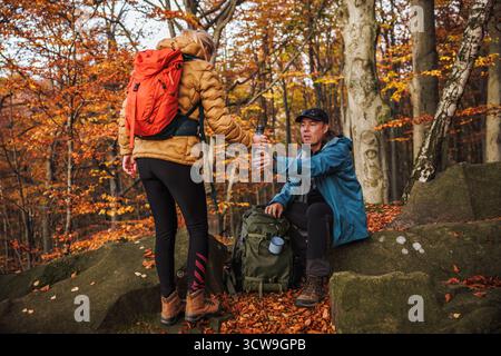 Zwei Wanderer ruhen sich nach einem erfolgreichen Wanderabenteuer im Herbstwald aus. Ein Tourist, der seiner Freundin Thermos mit heißem Getränk gibt Stockfoto