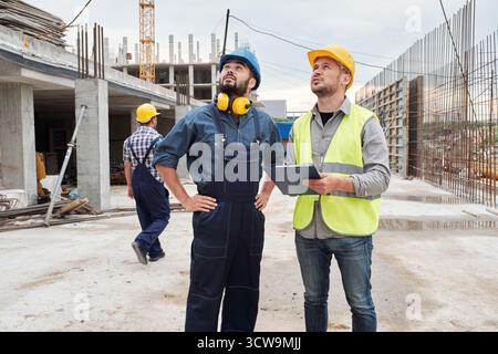 Junger Mann mit Sicherheitsweste und Helm, der eine Klemmplatte hält, der neben einem männlichen Kollegen mit Schutzhelm und Overall steht und die Baustelle im Freien inspiziert Stockfoto