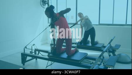Zwei Frauen in Sportbekleidung knieten bei Sonnenlicht im Pilates-Studio auf Reformern Stockfoto