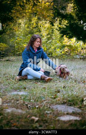 Frau streichelt und tröstet ihren Weimaranerhund, der im Herbst auf dem Gras auf einer sonnigen Waldlichtung liegt. Enge Verbundenheit, Loyalität und Gesellschaft Stockfoto