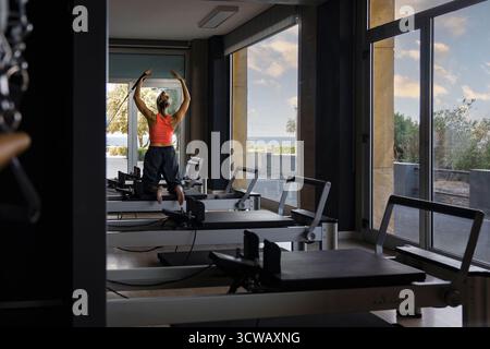 Eine nicht erkennbare athletische Frau von hinten hält eine Planke auf einem Pilates-Reformator und baut in einem modernen Fitnessstudio Stärke und Stabilität auf. Stockfoto