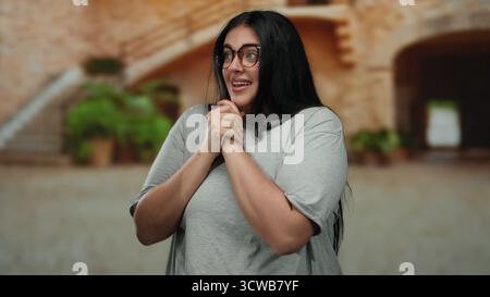 Die Frau, die im Freien in einer städtischen Straßenlage lächelt, zeigt Glück und Selbstvertrauen mit lässiger Kleidung und Brille, die einen charmanten Stil in Plus-Größe unterstreichen Stockfoto