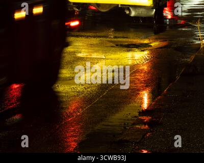 Regnerische nächtliche Stadtstraße mit nassem Asphalt und Wasser, das leuchtende gelbe und rote Fahrzeuglichter in eine verschwommene, abstrakte, leuchtende urbane Textur reflektiert Stockfoto