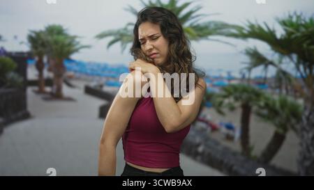 Frau in ärmellosem Oberteil, die Schulter neben Palmen auf der Strandstraße hält, während sie von Muskelanstrengung zuzwinkert; Schmerzen. Stockfoto