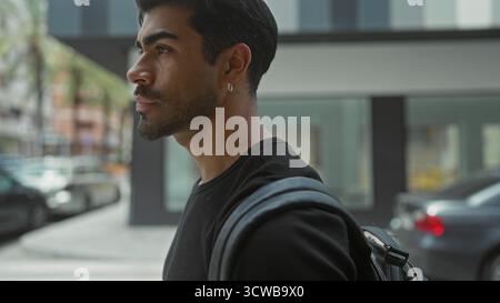 Mann in schwarzem Hemd mit Rucksack, der über die Schulter auf die Stadtstraße schaut, indem er eine Fassade inmitten verschwommener Autos baut; Neugier. Stockfoto