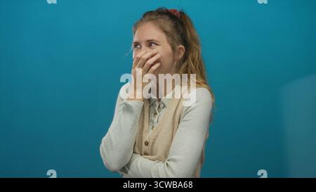 Die Frau, die die Hand benutzt, während sie auf einem Studio-Set lacht, zeigt einen blonden Stil und eine junge Atmosphäre, die einen blauen Hintergrund mit einer fröhlichen Lächelgeste bedeckt. Stockfoto