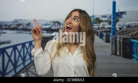 Die junge blonde Frau in weißem Hemd zeigt den Finger nach oben auf den hölzernen Pier am Yachthafen; aufregende Überraschung. Stockfoto