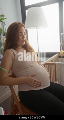 Junge kaukasische Frau, die auf Holzstuhl sitzt und schwangeren Bauch hält und im Kinderzimmer grimmt; Unwohlsein. Stockfoto