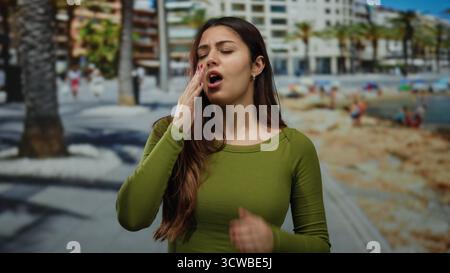 Die junge Frau, die am Strand niest, sieht unter sonnigem Himmel mit verschwommenen Palmen im Hintergrund unwohl aus. Stockfoto