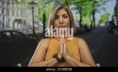 Junge blonde Frau mit gefalteten Händen auf der Brust steht inmitten geparkter Autos auf belebter, sonnendurchfluteter Stadtstraße; Gelassenheit. Stockfoto