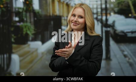 Frau klatscht auf der Straße der Stadt die Hände, zeigt Jugendliche Energie und professionelle Kleidung in urbaner Umgebung mit blondem Haar, was eine selbstbewusste und stilvolle Atmosphäre schafft Stockfoto