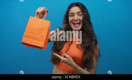 Die junge hispanische Frau lächelt und zeigt den Finger auf die orangefarbene Einkaufstasche im Studio; Aufregung. Stockfoto