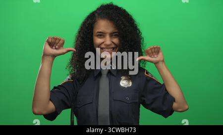 Hispanische Offizierin in Uniform lächelt selbstbewusst mit Daumen, die vor einer grünen Hintergrundwand auf sich gerichtet sind. Stockfoto