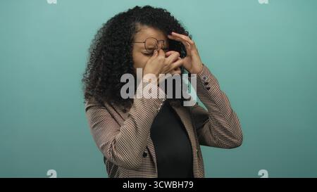 Eine Frau mit lockigem Haar, die Brille und brauner Jacke trägt und ihren Kopf auf isoliertem grünen Hintergrund hält, ruft Stress oder Kopfschmerzen hervor. Stockfoto