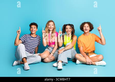 Vier Teenager lachen und zeigen, während sie zusammen in einer bunten, lässigen Street-Style-Szene vor einem leuchtend blauen Hintergrund sitzen Stockfoto
