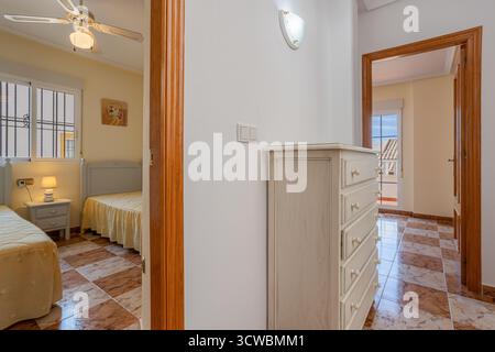 Heller Flur mit weißer Kommode, Zugang zu einem Zweibettzimmer mit Deckenventilator und einem zweiten Zimmer mit Balkon und Blick auf den Außenbereich. Stockfoto