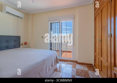 Sonniges Schlafzimmer mit großem Bett mit gepolstertem Kopfteil, eingebautem Holzschrank, Klimaanlage und Glasschiebetüren zu einem privaten Balkon. Stockfoto