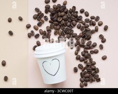 Geröstete Kaffeebohnen, die auf einem zweifarbigen Hintergrund in der Nähe einer Papiertasse verstreut sind, auf der ein Herz gezeichnet ist, was die Liebe zum Kaffee zum Ausdruck bringt Stockfoto