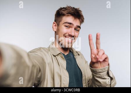 Porträt eines jungen Mannes, der ein fröhliches Selfie mit einem Friedenszeichen macht, das Freude und spielerische Energie auf weißem Hintergrund ausstrahlt Stockfoto
