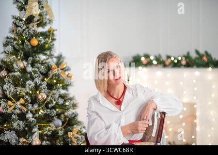 Stilvolle Frau mit blonden Haaren sitzt in einem gemütlichen Stuhl, umgeben von festlichen Dekorationen und einem leuchtenden Weihnachtsbaum, was an einen fröhlichen Feiertag erinnert Stockfoto