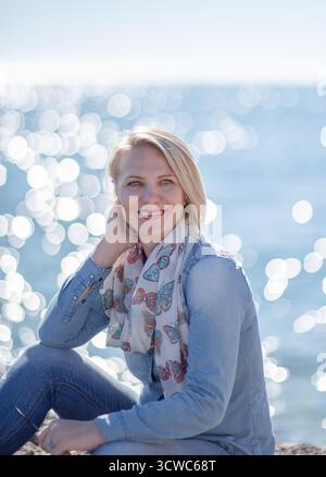 Die blonde Frau sitzt am Meer und trägt ein Jeansshirt und einen Schal, wobei das Sonnenlicht auf dem Wasser reflektiert und eine fröhliche und ruhige Atmosphäre schafft Stockfoto