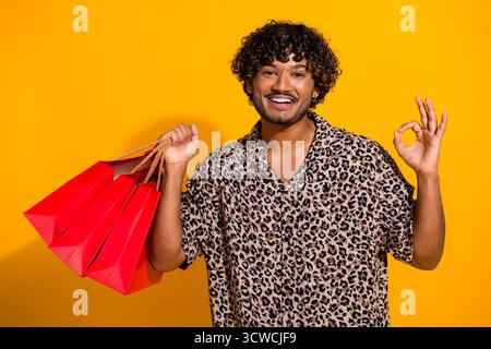 Fröhlicher junger Mann, der rote Einkaufstaschen hält, während er eine gute Geste vor einem leuchtend gelben Hintergrund macht Stockfoto