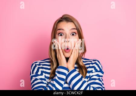 Überraschte junge Frau in lässigem gestreiftem Pullover, posiert vor rosa Hintergrund mit einer fröhlichen und ausdrucksstarken Reaktion Stockfoto