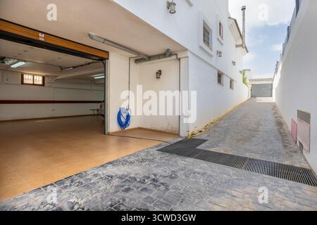 Außenansicht eines Garageneingangs in einem Wohnhaus mit offener Sektionaltür, die den Innenparkplatz freigibt. Die schräge Auffahrt ist mit Stein gepflastert Stockfoto