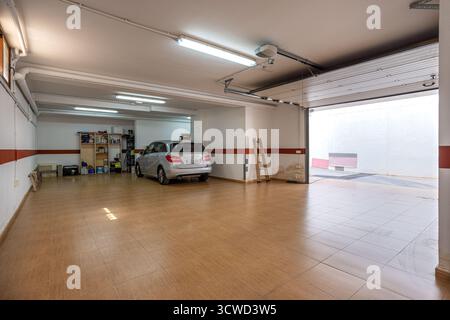 Große und saubere Wohngarage mit einem geparkten silbernen Auto, Fliesenboden und einem teilweise offenen Garagentor. Der Raum ist gut beleuchtet wi-fi Stockfoto