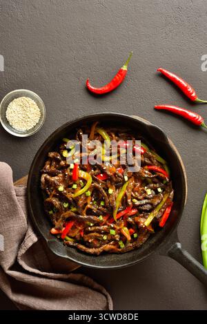 Frittiertes, dünn geschnittenes Rindersteak mit Paprika in gusseiserner Pfanne auf dunkelbraunem Hintergrund. Draufsicht, flach Stockfoto