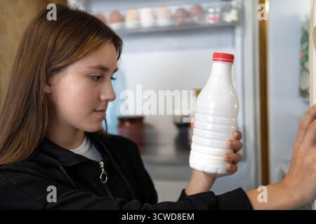 Hungriges Mädchen macht leeren Kühlschrank auf. Ein Teenager-Mädchen sucht im Kühlschrank nach Essen und wählt aus, was er essen soll. Das Mädchen nimmt eine weiße Flasche frische Milch Stockfoto