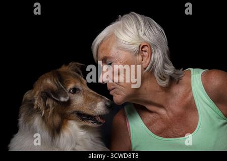 Ein herzerwärmendes Atelier-Bild einer älteren Frau, die sich sanft zu ihrem Collienhund für einen Kuss beugt. Das Foto wird vor schwarzem Hintergrund aufgenommen Stockfoto