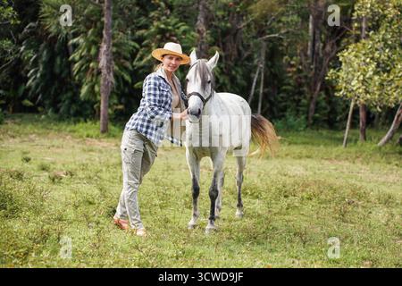 Junge Frau in Hemd und Strohhut posiert neben weißem grauem Pferd, Dschungel-Bäume Hintergrund - Reiten, Ranch im Isalo Park, Madagaskar Stockfoto