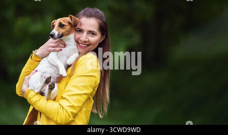 Junge Frau in gelber Jacke, die ihren Jack Russell Terrier Hund an den Händen hält, verschwommener grüner Baumhintergrund Stockfoto