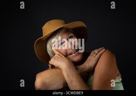 Ein fröhliches und herzerwärmendes Studio-Porträt einer älteren Frau mit Strohsonnenhut. Sie lehnt ihren Kopf mit einem hellen Lächeln auf ihre Hand und schafft einen Stockfoto
