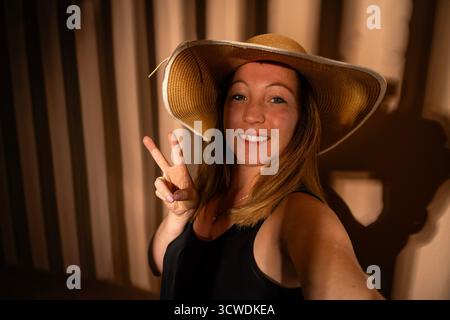 Eine junge Frau mit Strohhut und schwarzem Oberteil, lächelt und macht eine Friedenszeichen-Geste Stockfoto