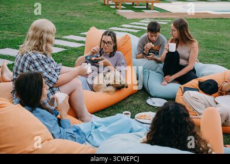 Freunde, die sich auf bunten Sitzsäcken im Freien entspannen und Essen und Getränke in zwangloser geselliger Atmosphäre mit einem kleinen Hund teilen. Frau informell Stockfoto