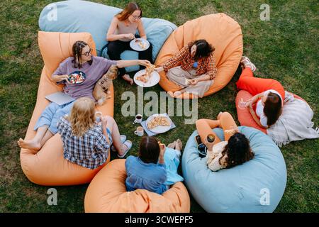 Freunde, die sich auf bunten Sitzsäcken im Freien entspannen und Essen und Getränke in zwangloser geselliger Atmosphäre mit einem kleinen Hund teilen. Frau informell Stockfoto