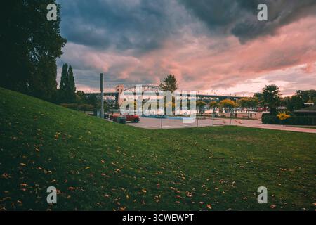 Malerischer Blick auf einen Fluss bei Sonnenuntergang mit dunklen Wolken darüber. Eine Brücke überspannt das Wasser, umgeben von üppigem Grün und lebhaften Blumen. Stockfoto