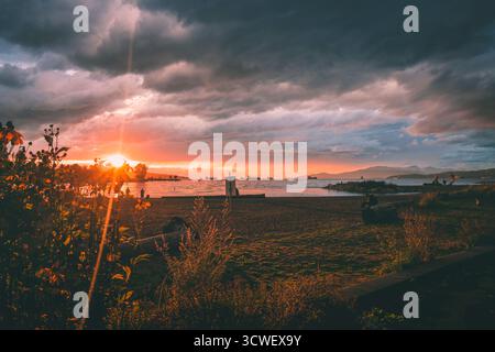 Der wunderschöne Sonnenuntergang beleuchtet die Bucht mit Silhouetten von Schiffen, während die Menschen die Aussicht entlang der Küste genießen. Stockfoto