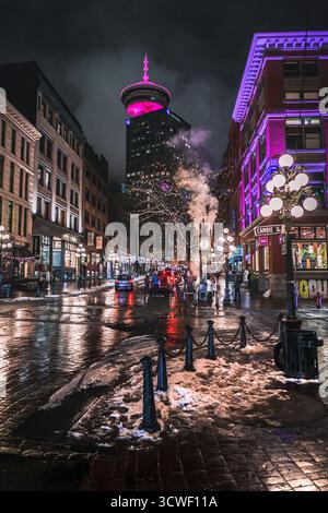 Die Menschen schlendern nachts durch eine lebhafte Stadtstraße, mit bunten Lichtern, die auf nassem Gehweg reflektieren, und einem Turm, der darüber leuchtet. Stockfoto