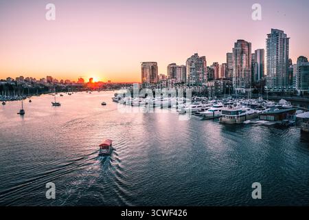 Ein Boot segelt im Hafen, während die Sonne hinter den Gebäuden der Stadt untergeht und warme Farben über das Wasser und die Skyline wirft. Stockfoto