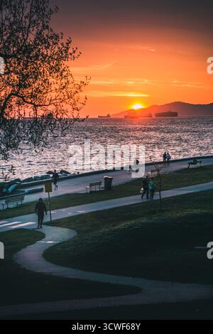 Die Menschen laufen entlang des Uferwegs, während die Sonne über der Bucht untergeht und warme Farben über den Himmel und das Wasser wirft. Stockfoto