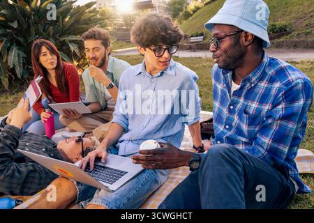 Eine Gruppe fröhlicher junger multiethnischer Studenten arbeitet an einem Projekt mit einem Laptop und Tablets zusammen, während sie gemeinsam auf einer Decke in einem Park Dura studieren Stockfoto