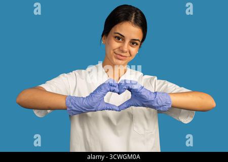 Junge weibliche Gesundheitsberaterin, die eine Herzform mit Handschuhen vor blauem Hintergrund macht Stockfoto