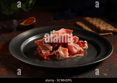 Teller mit köstlichen Schinkenscheiben und Knäckebrot auf dunklem Tisch Stockfoto